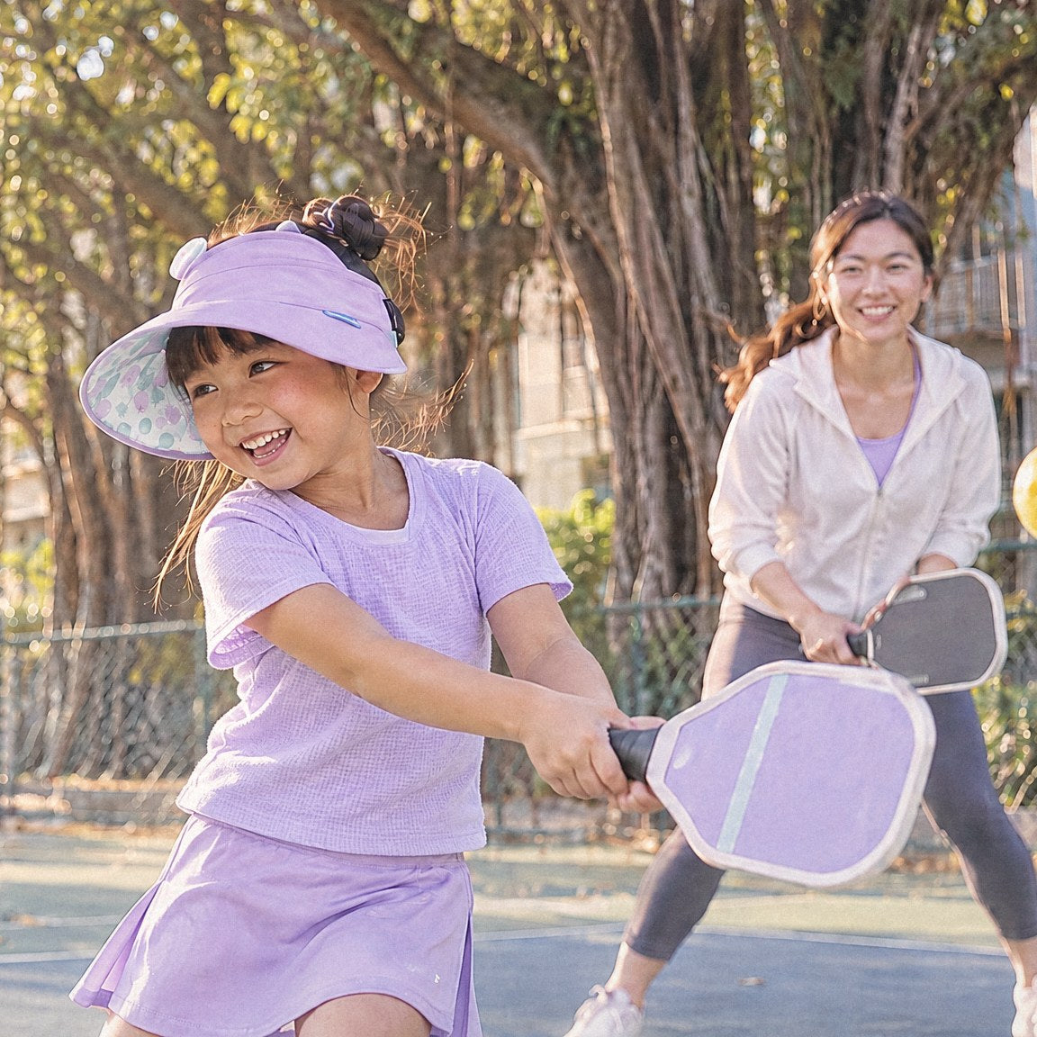 穿淡紫色上衣的女孩戴泡泡水球雙面中空遮陽帽，和媽媽在公園球場打匹克球